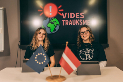 Jaunie vides reportieri publicitātes foto. Divas jaunietes - raidījuma vadītājas - sēž ar uzrakstu "Vides trauksme" fonā