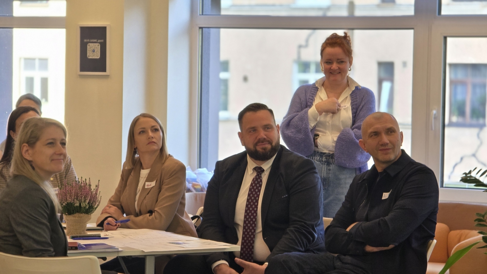 Darbnīcas dalībnieki sēž ap darba galdu un klausās prezentāciju