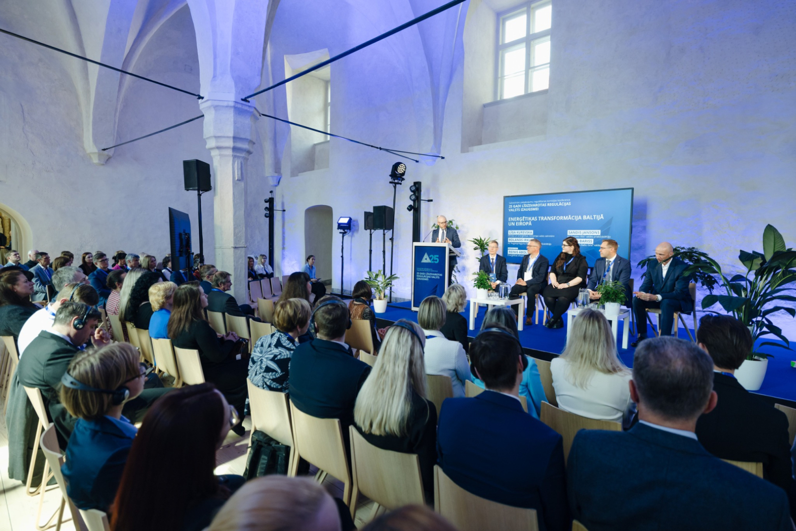 Zāle ar konferences salībniekiem, kas klausās diskusiju uz skatuves