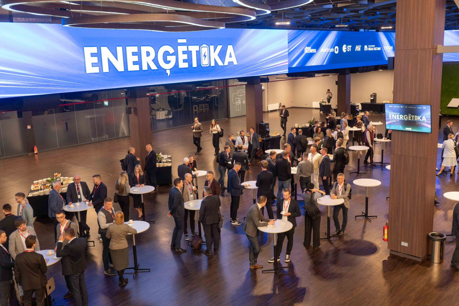 Zāle ar konferences apmeklētājiem, kas stāv pie stāvgaldiem. Fonā redzams uzraksts "Enerģētika"