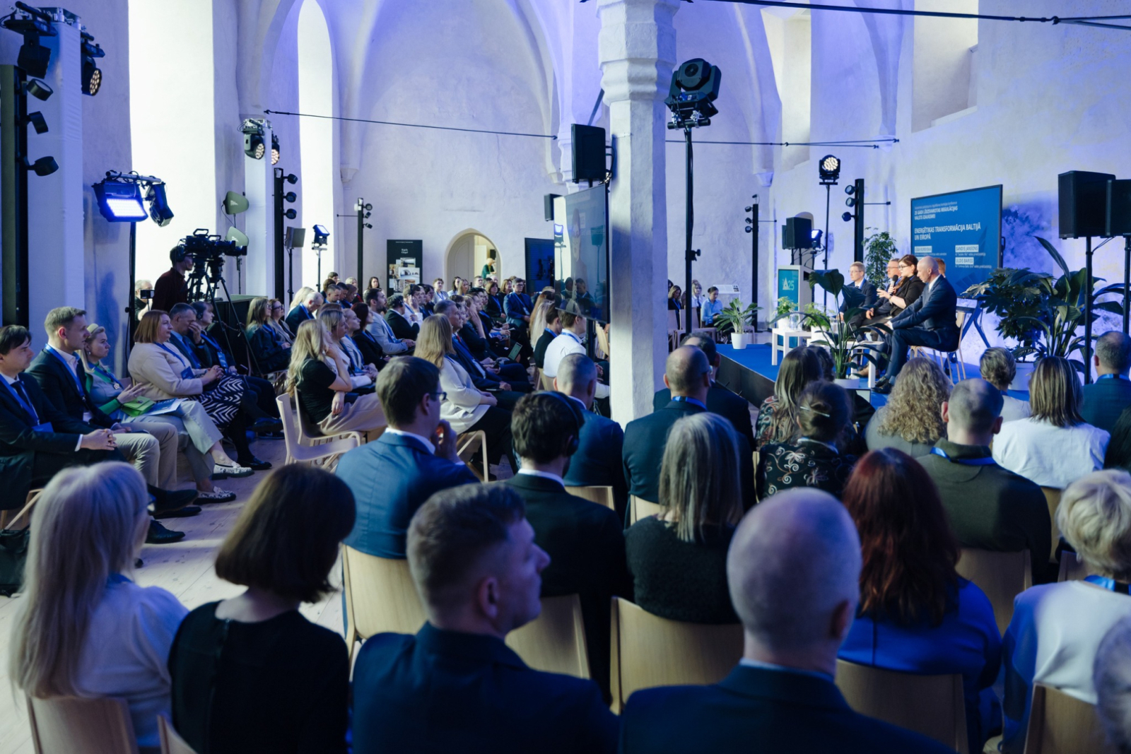 Zāle ar konferences salībniekiem, kas klausās diskusiju uz skatuves