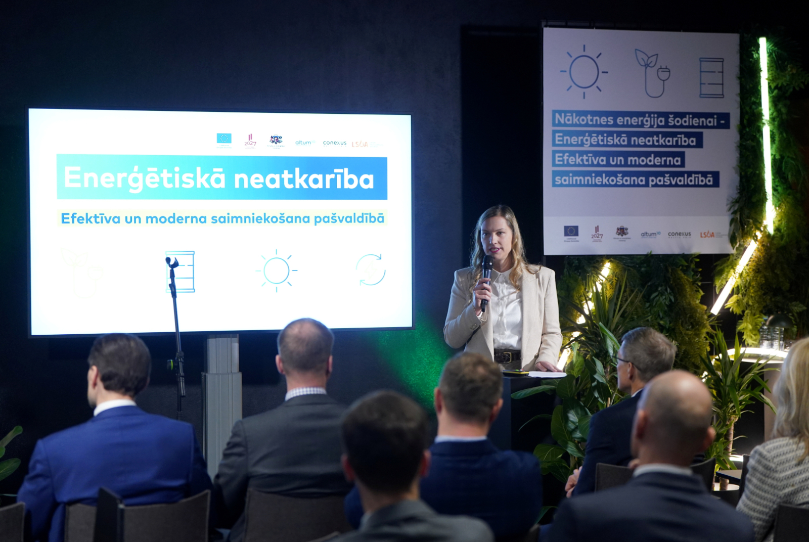 Uzruna zāles priekšā ar fonā redzamu uzrakstu "Enerģētiskā neatkarība. Efektīva un moderna saimniekošana pašvaldībā"