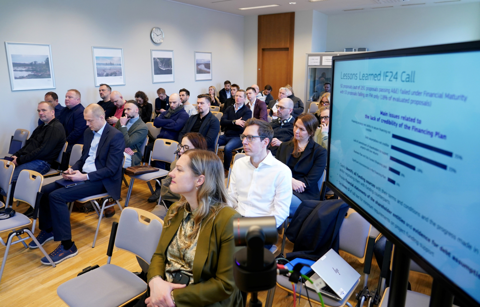 Zāle, kurā sēž info dienas dalībnieki un ekrāns ar prezentāciju