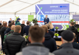 Uzruna uz skatuves pasākuma atklāšanā zālei ar sēdošiem dalībniekiem